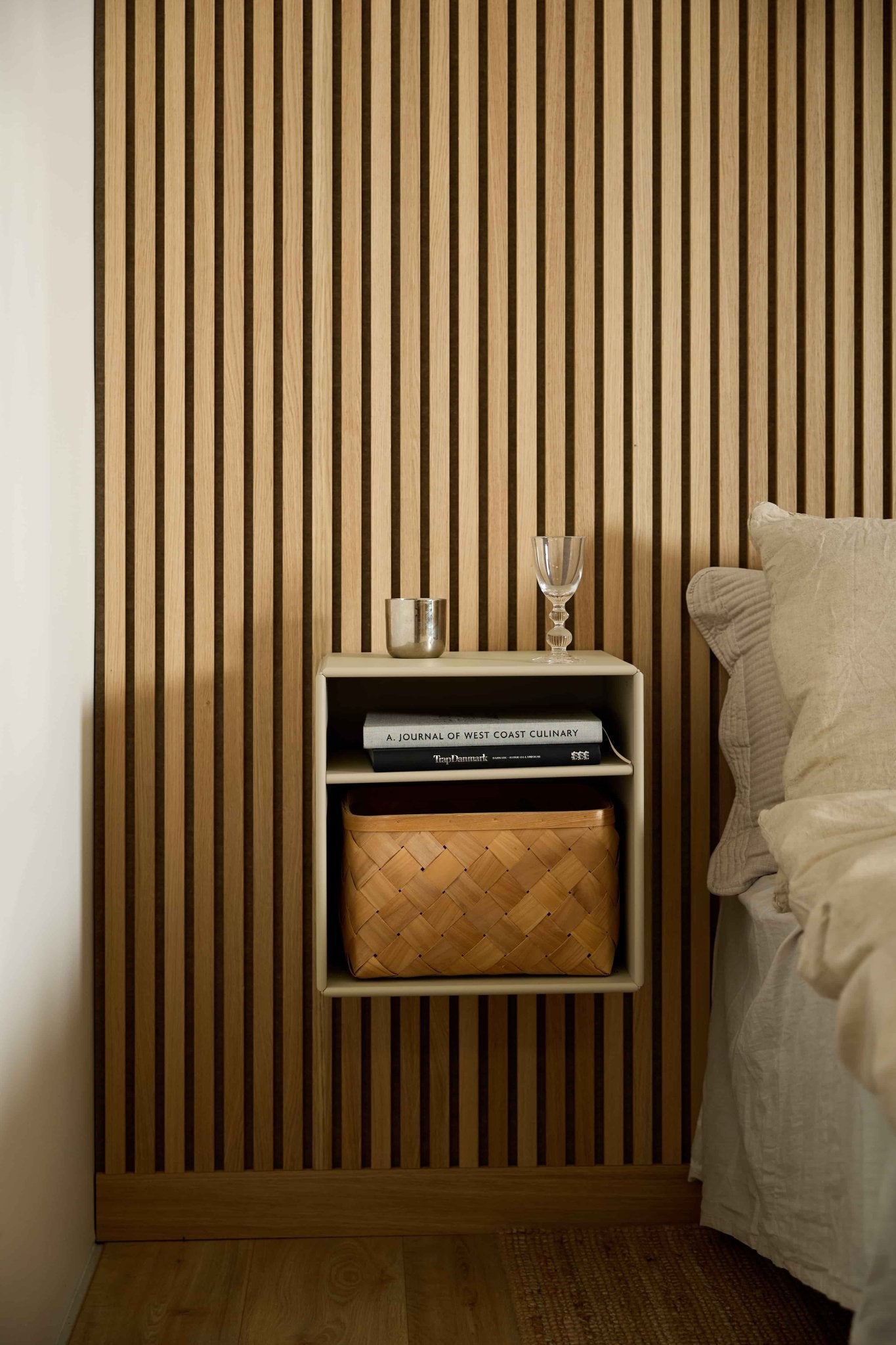 Estante WoodUpp Arka em bege ao lado de uma cama, com fundo numa parede acústica de ripas de madeira Akupanel, decorada com livros e elementos minimalistas.